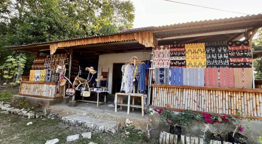 A souvenir shop with a corrugated roof and bamboo railing displays colorful Sumba ikat textiles, clothing, hats, and bags. Green trees are in the background, with pink flowers in the foreground.