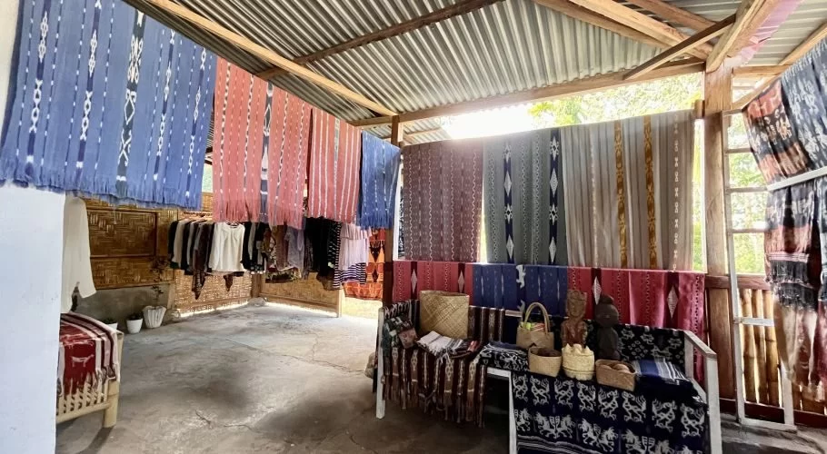 Colorful Sumba ikat textiles with intricate patterns hang from wooden beams in an open-air shop, with more fabrics and crafts displayed on a bench.