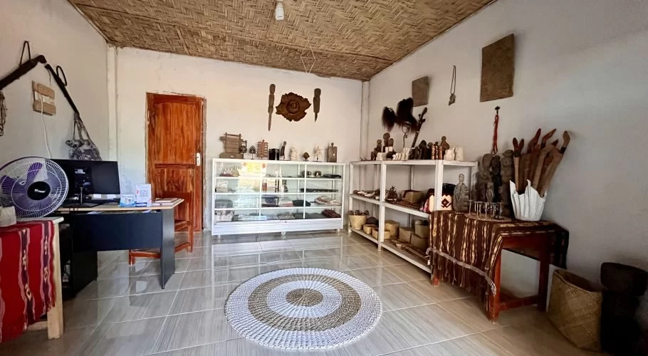A bright souvenir shop interior features white walls, a woven ceiling, and tiled floor, displaying Sumba textiles, carvings, and crafts on shelves and a desk.
