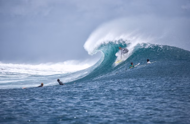 A surfer in a grey wetsuit rides a large, turquoise-blue breaking wave on a yellow board, with other surfers waiting in the deep blue ocean under a cloudy sky.