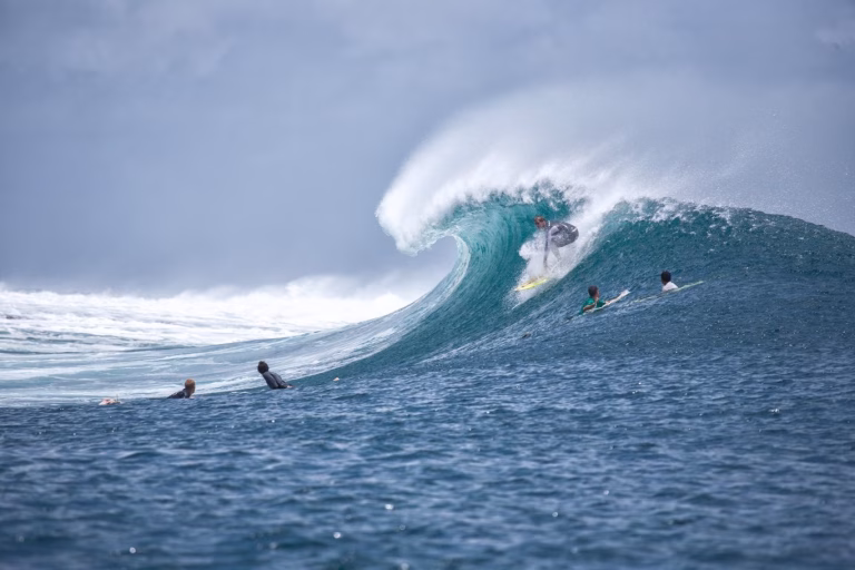 A surfer in a grey wetsuit rides a large, turquoise-blue breaking wave on a yellow board, with other surfers waiting in the deep blue ocean under a cloudy sky.