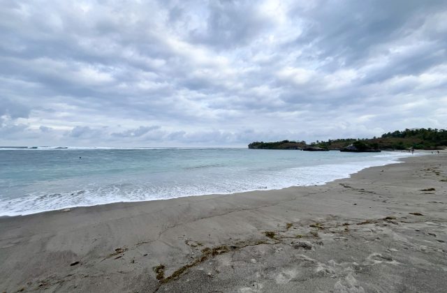 A wide view of Kerewe Beach shows grey sand, blue-green ocean with breaking waves, a rocky headland, and a cloudy sky.