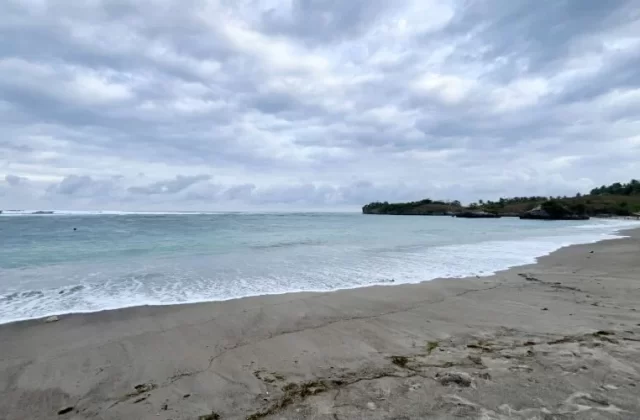 A wide view of Kerewe Beach shows grey sand, blue-green ocean with breaking waves, a rocky headland, and a cloudy sky.