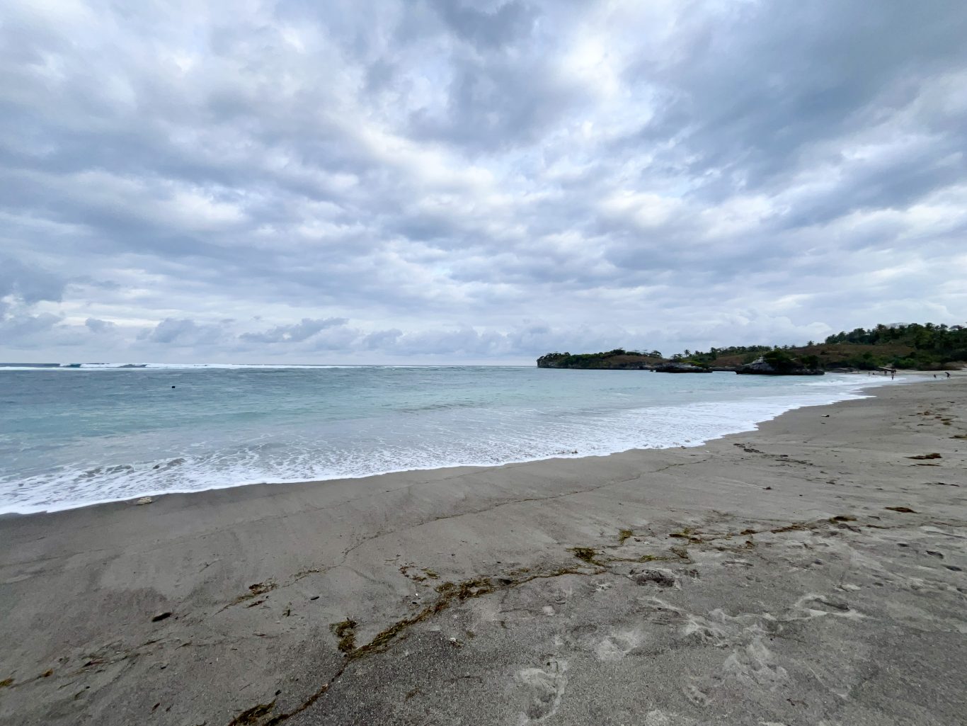A wide view of Kerewe Beach shows grey sand, blue-green ocean with breaking waves, a rocky headland, and a cloudy sky.