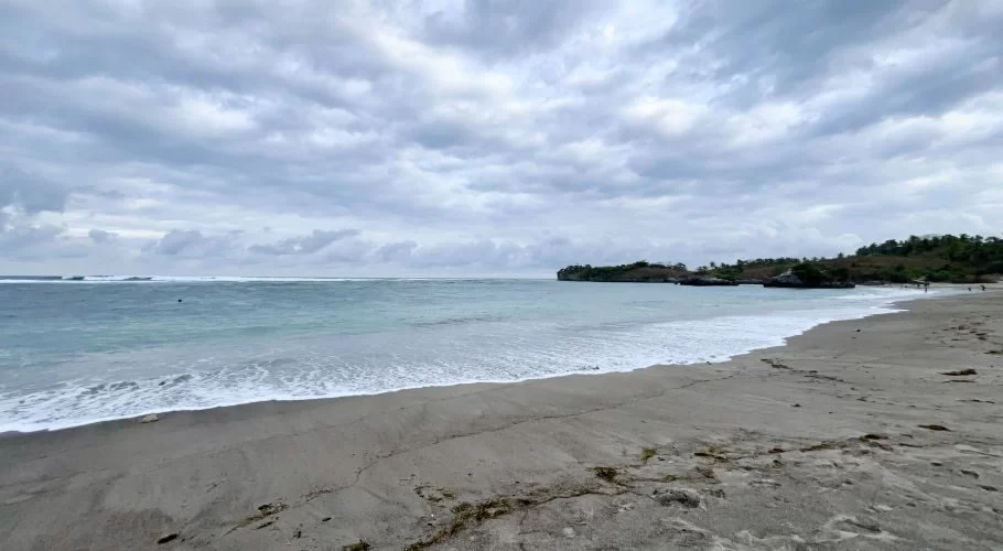 A wide view of Kerewe Beach shows grey sand, blue-green ocean with breaking waves, a rocky headland, and a cloudy sky.