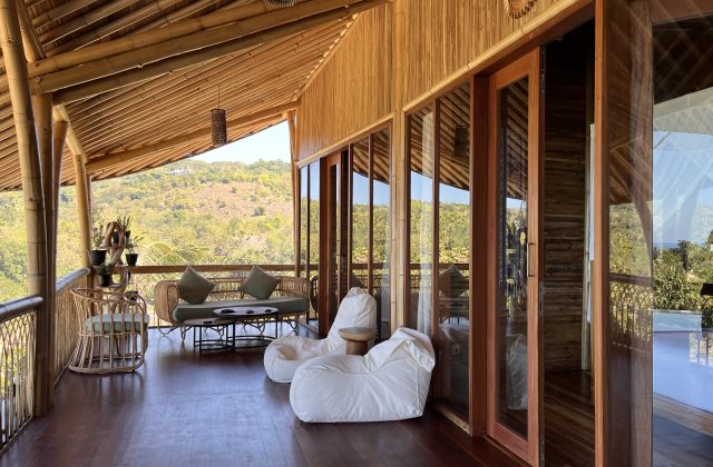 A bamboo lodge balcony features rattan seating, white bean bags, and glass doors, offering views of green hills and a distant ocean.