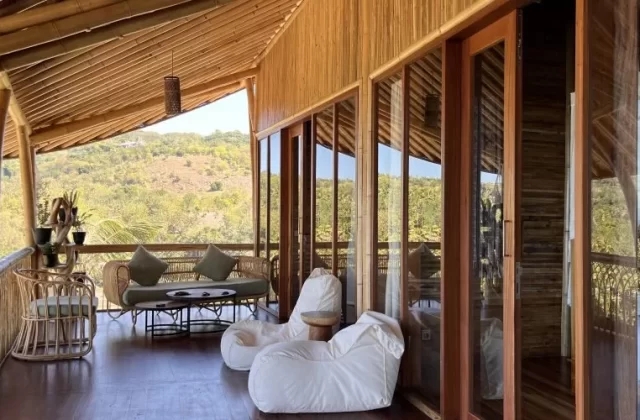 A bamboo lodge balcony features rattan seating, white bean bags, and glass doors, offering views of green hills and a distant ocean.