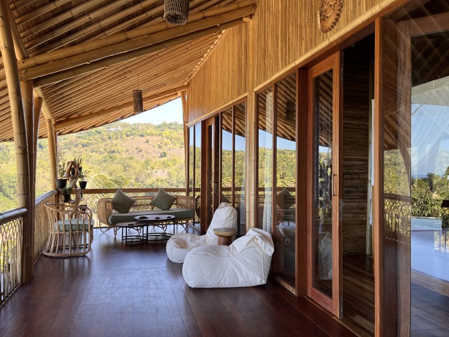 A bamboo lodge balcony features rattan seating, white bean bags, and glass doors, offering views of green hills and a distant ocean.
