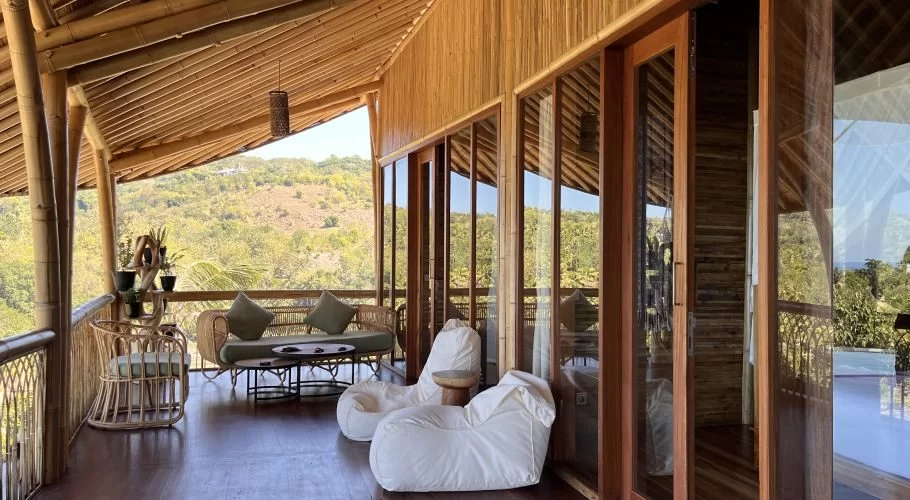 A bamboo lodge balcony features rattan seating, white bean bags, and glass doors, offering views of green hills and a distant ocean.