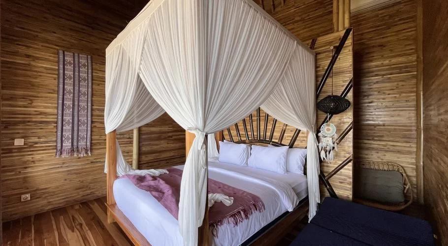 A spacious bedroom in a bamboo lodge features a four-poster bed with white netting, white bedding, and a purple throw, set against light brown wooden walls.