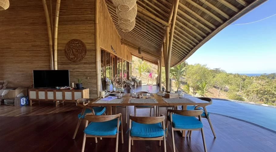 A spacious open-air dining area at Ketanu Sumba Bamboo Lodge features a long wooden table with blue chairs, overlooking an infinity pool, lush trees, and the ocean under a clear sky.