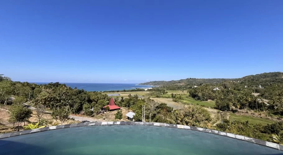 A vibrant blue infinity pool overlooks a lush green landscape with a traditional red-roofed Sumba house, distant blue ocean, white beach, and rolling hills under a clear sky.