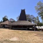 A traditional Sumbanese house with a tall, conical thatched roof stands on dry ground, flanked by an Indonesian flag and other village buildings under a clear blue sky.