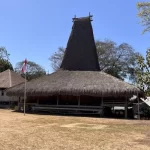 A traditional Sumbanese house with a tall, conical thatched roof stands on dry ground, flanked by an Indonesian flag and other village buildings under a clear blue sky.