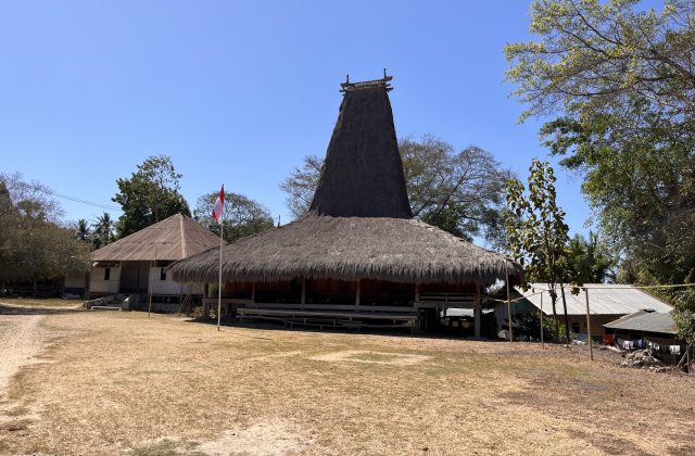 A traditional Sumbanese house with a tall, conical thatched roof stands on dry ground, flanked by an Indonesian flag and other village buildings under a clear blue sky.