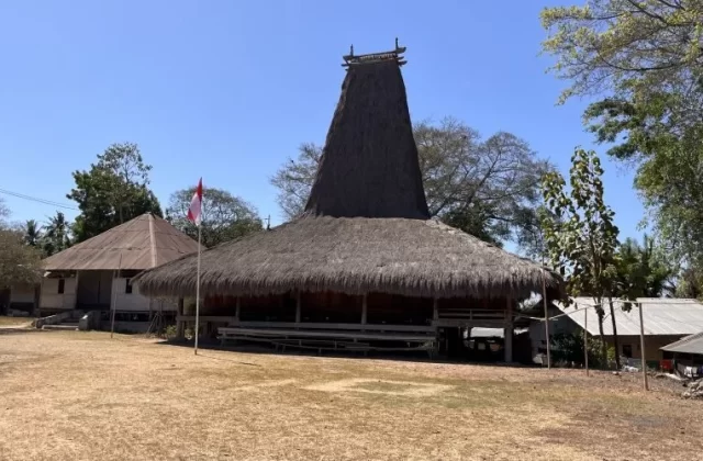 A traditional Sumbanese house with a tall, conical thatched roof stands on dry ground, flanked by an Indonesian flag and other village buildings under a clear blue sky.