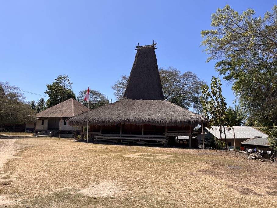 A traditional Sumbanese house with a tall, conical thatched roof stands on dry ground, flanked by an Indonesian flag and other village buildings under a clear blue sky.
