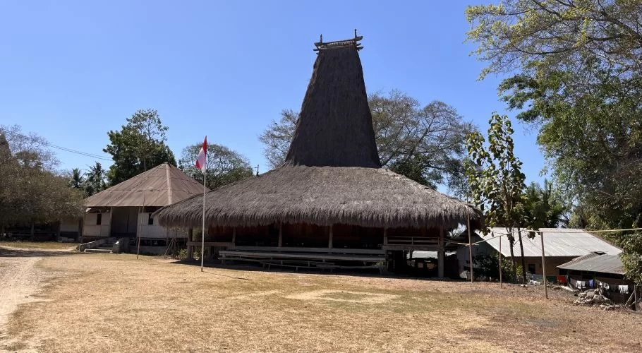 A traditional Sumbanese house with a tall, conical thatched roof stands on dry ground, flanked by an Indonesian flag and other village buildings under a clear blue sky.