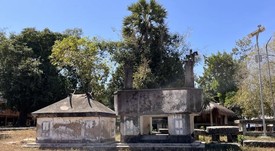 Visible are two weathered grey megalithic graves, one with a horned roof, under a clear blue sky amidst green trees and dry grass.