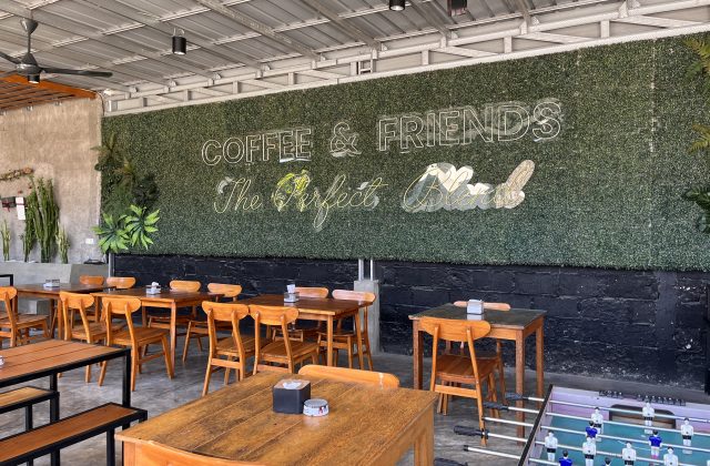 A modern cafe interior features numerous wooden tables and chairs, a green artificial hedge wall with neon signs, and an industrial ceiling.