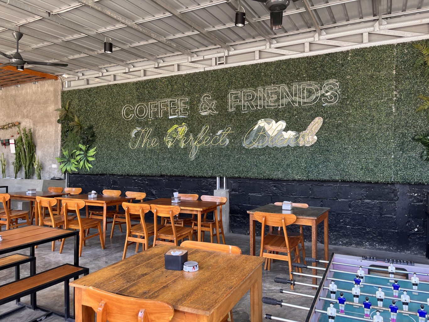 A modern cafe interior features numerous wooden tables and chairs, a green artificial hedge wall with neon signs, and an industrial ceiling.