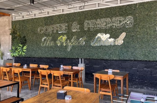 A modern cafe interior features numerous wooden tables and chairs, a green artificial hedge wall with neon signs, and an industrial ceiling.