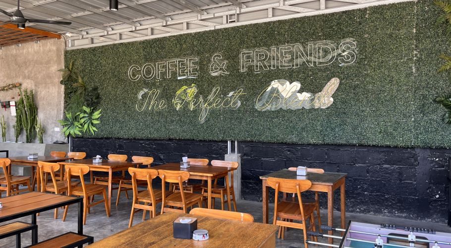 A modern cafe interior features numerous wooden tables and chairs, a green artificial hedge wall with neon signs, and an industrial ceiling.