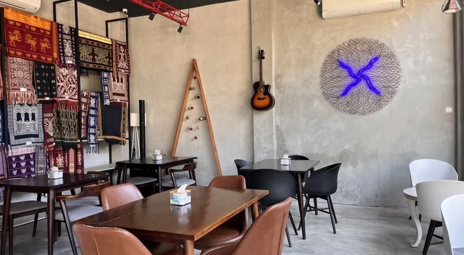 Modern cafe interior with concrete walls, dark tables, and chairs. Colorful Sumba ikat textiles, a guitar, and a glowing blue art piece.