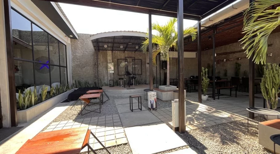 A modern outdoor cafe features wooden tables, gravel paths, and a stage with musical instruments, all under a partial roof with bright sunlight.