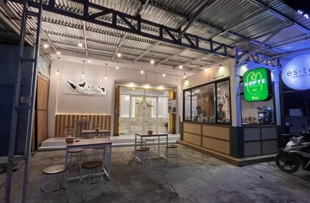 A brightly lit modern cafe exterior at night in Waingapu, Sumba, features outdoor seating, a "Kopte" sign, and a scooter parked nearby.