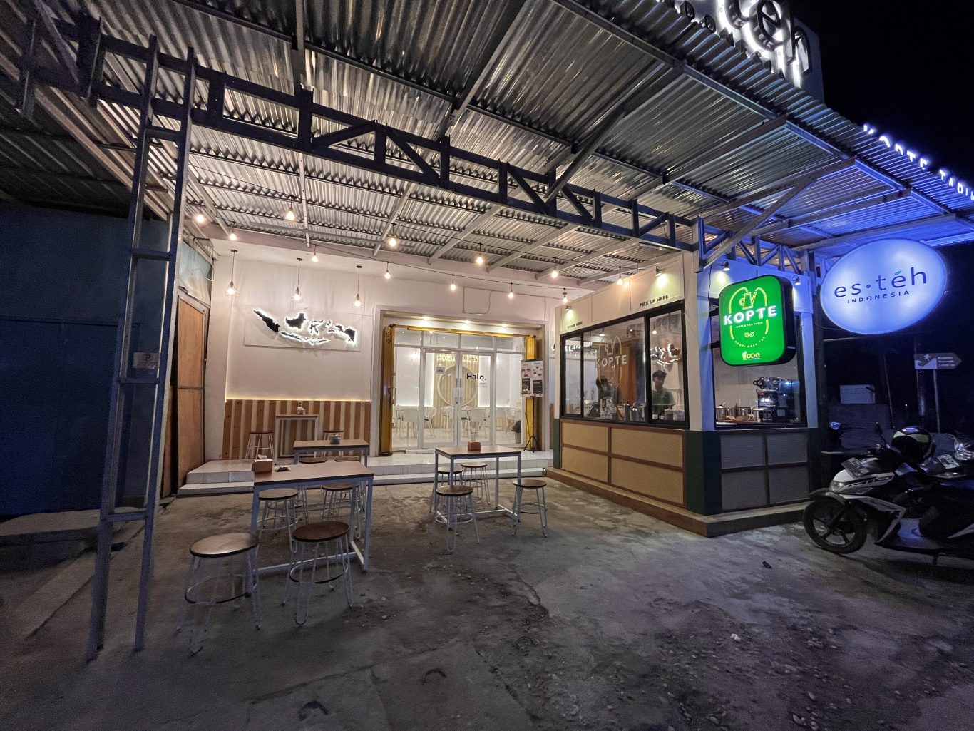 A brightly lit modern cafe exterior at night in Waingapu, Sumba, features outdoor seating, a "Kopte" sign, and a scooter parked nearby.