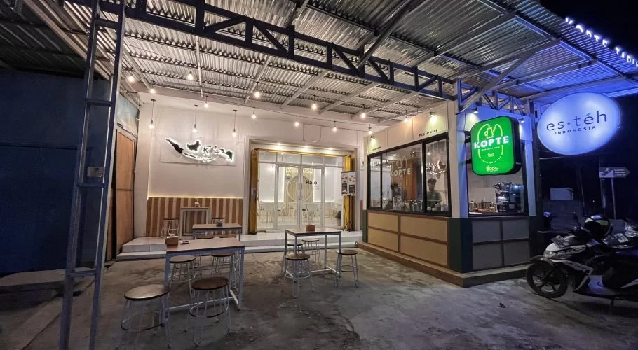 A brightly lit modern cafe exterior at night in Waingapu, Sumba, features outdoor seating, a "Kopte" sign, and a scooter parked nearby.