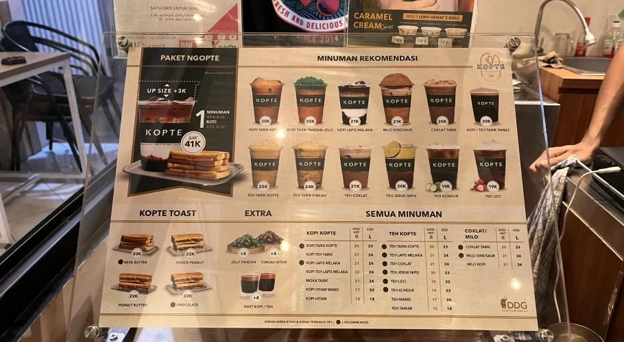 A brightly lit menu board for Kopte cafe displays various modern coffee, tea, and toast options with prices, set within a contemporary interior.