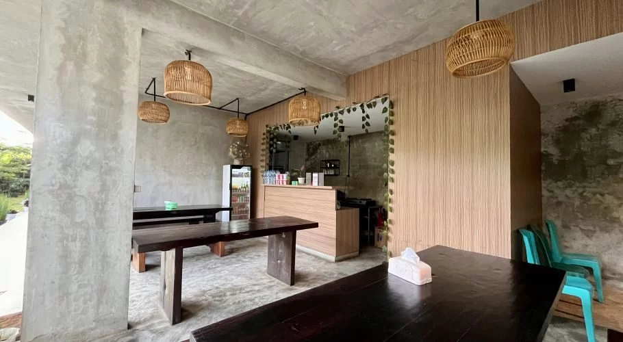 A minimalist cafe interior with concrete walls, dark wooden tables, and rattan pendant lights, featuring a light wood counter and a glimpse of a green garden outside.