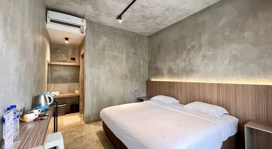 A minimalist hotel room features a large bed with white linens, a wooden headboard with backlighting, concrete walls, an air conditioner, and a desk with a kettle.