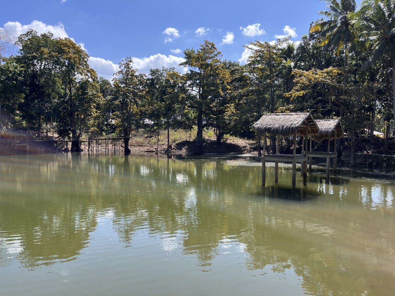 A serene green lake in Sumba is surrounded by lush trees under a blue sky, featuring two thatched gazebos on stilts over the water.