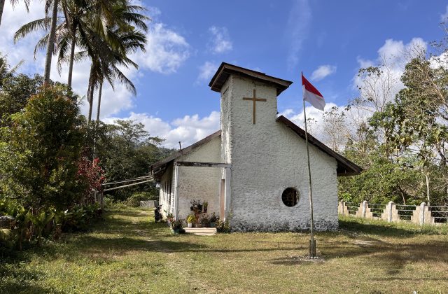 Lakoka Church, an old white stone building with a cross and a round window, stands under a blue sky with an Indonesian flag, surrounded by green trees.