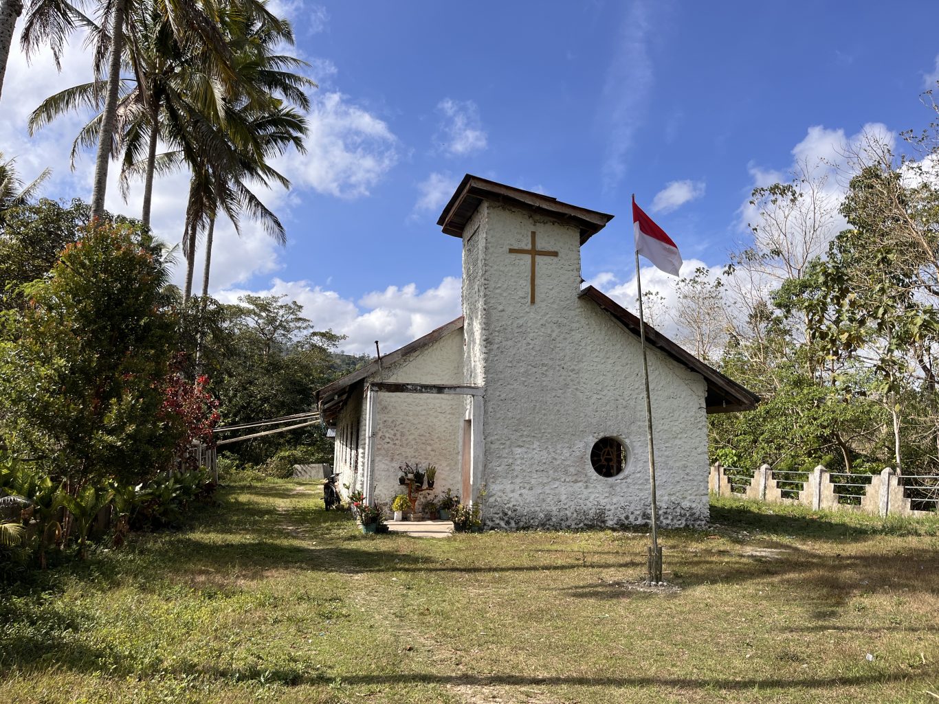 Lakoka Church, an old white stone building with a cross and a round window, stands under a blue sky with an Indonesian flag, surrounded by green trees.