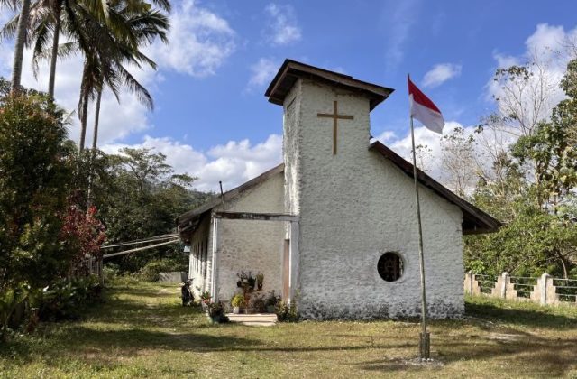 Lakoka Church, an old white stone building with a cross and a round window, stands under a blue sky with an Indonesian flag, surrounded by green trees.