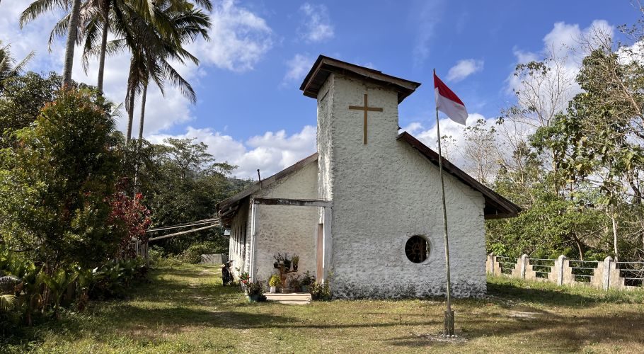 Lakoka Church, an old white stone building with a cross and a round window, stands under a blue sky with an Indonesian flag, surrounded by green trees.