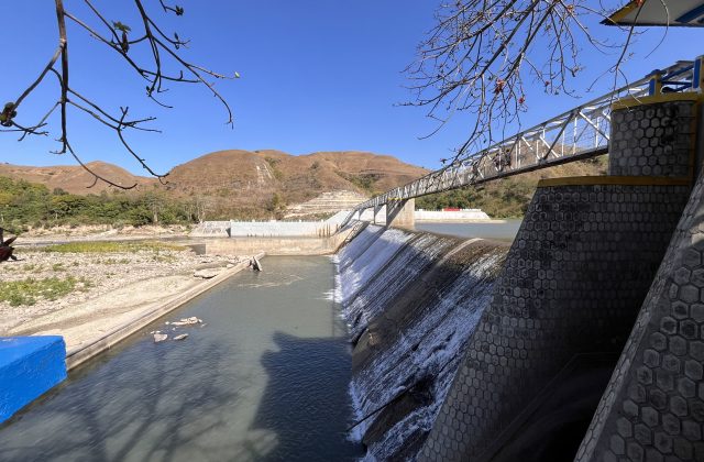 Lambanapu Dam in East Sumba features water cascading over concrete walls, a pedestrian bridge with people, and dry brown hills under a clear blue sky.