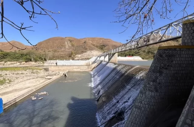 Lambanapu Dam in East Sumba features water cascading over concrete walls, a pedestrian bridge with people, and dry brown hills under a clear blue sky.