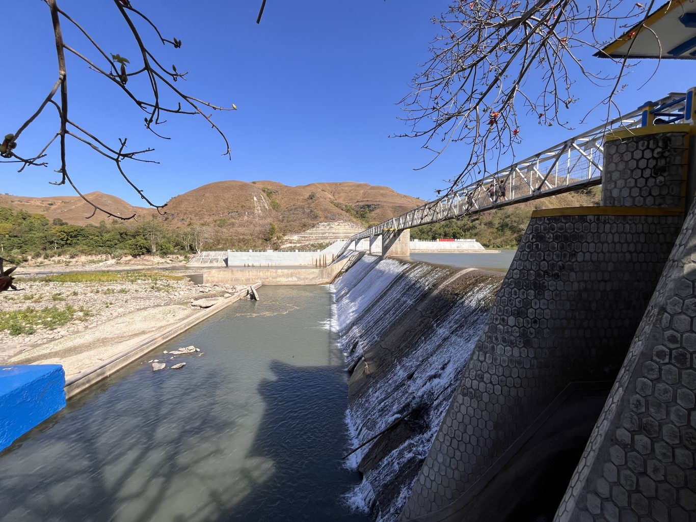 Lambanapu Dam in East Sumba features water cascading over concrete walls, a pedestrian bridge with people, and dry brown hills under a clear blue sky.