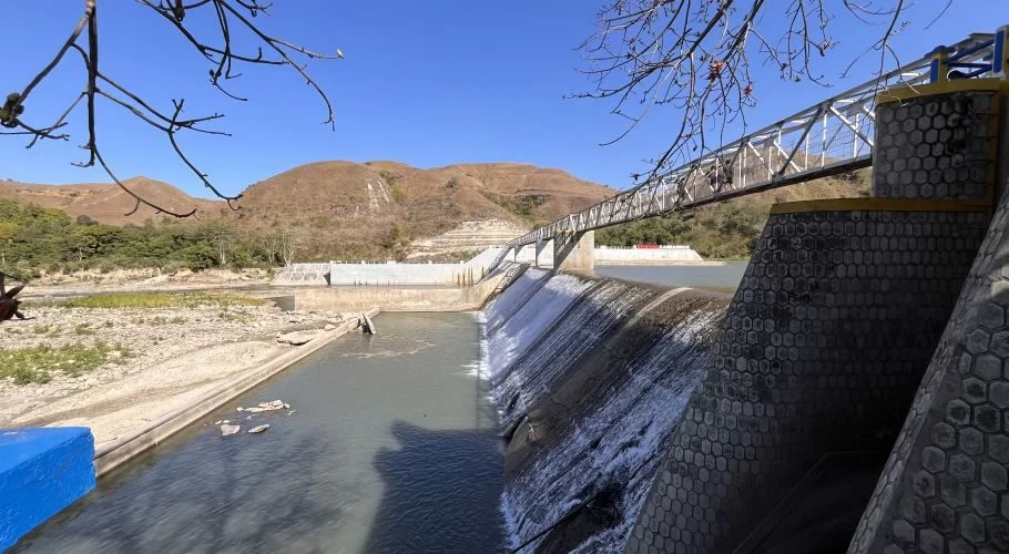 Lambanapu Dam in East Sumba features water cascading over concrete walls, a pedestrian bridge with people, and dry brown hills under a clear blue sky.