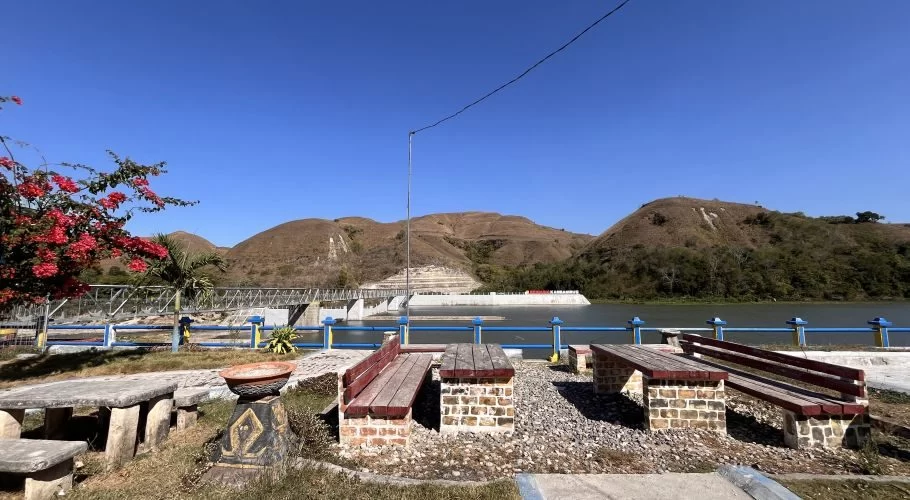 Seating areas with stone tables and wooden benches in the foreground, overlooking the Lambanapu Dam and its reservoir with a bridge, backed by dry, rolling hills under a clear blue sky. Red flowers are on the left.