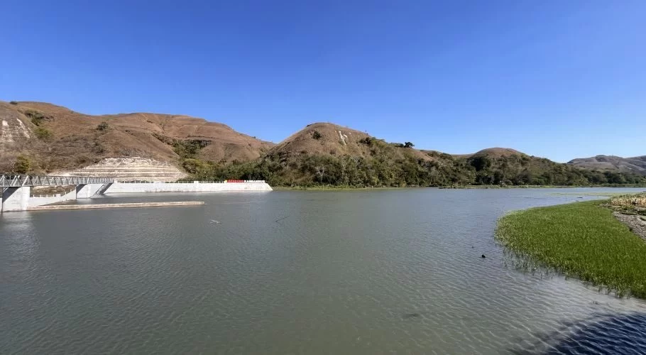 Lambanapu Dam's reservoir with a concrete dam structure and bridge on the left. Rolling brown hills rise under a clear blue sky, and green reeds line the water's edge.