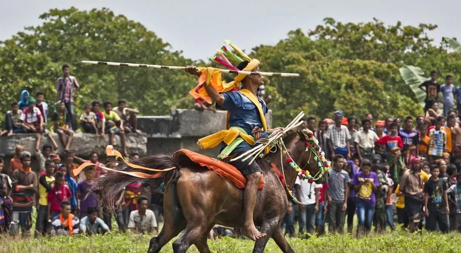 A Sumbanese man on a brown horse, adorned in colorful traditional attire, holds a wooden spear high, with a crowd and green trees.