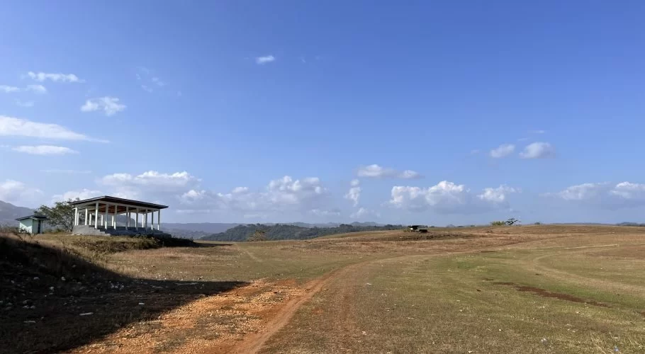 A white open-sided pavilion stands on a grassy hill overlooking a vast dry field with a dirt path, under a bright blue sky with white clouds.