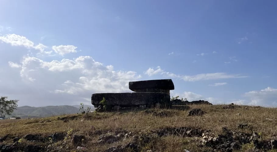 Two dark, ancient megalithic tomb stones sit on a dry, grassy hill under a bright blue sky with white clouds.