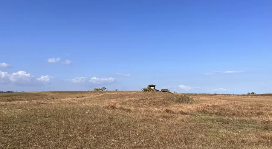 A wide, dry, golden-brown field under a bright blue sky with scattered clouds. Two dark megalithic tombs are visible on a distant rise.
